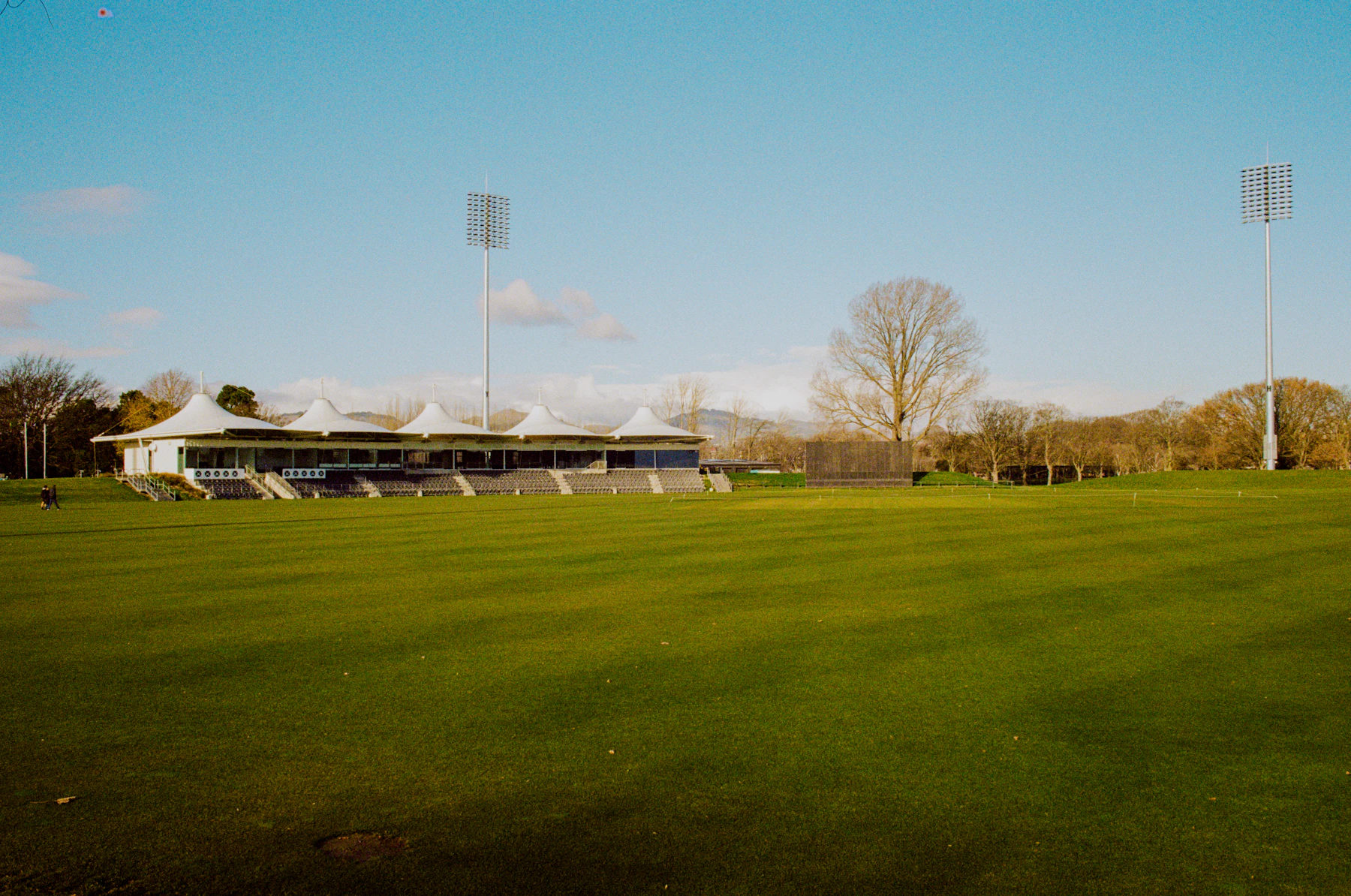A village cricket ground under a pale sky, a small pavilion at the far boundary