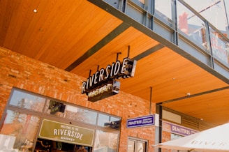 A storefront with a signboard reading 'Riverside Market' is visible, set against a building with brick walls and a wooden roof overhang. Other signs, including 'Dimitris Greek Food Souvlaki', can be seen nearby. There is a modern architectural style with metal and glass elements.