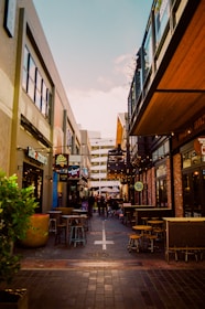 A vibrant urban alleyway with a variety of shops and cafes along both sides. Outdoor seating is available in the form of small tables and stools, and string lights hang above, adding a warm atmosphere. The alley is lined with people walking in the distance.