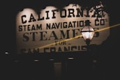 A vintage-style signboard featuring bold, black lettering with the words 'California Steam Navigation Co's Steamers for San Francis'. A lamp post with a lit lantern is prominently in the foreground, casting a warm glow against the dimly lit surroundings.