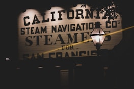 A vintage-style signboard featuring bold, black lettering with the words 'California Steam Navigation Co's Steamers for San Francis'. A lamp post with a lit lantern is prominently in the foreground, casting a warm glow against the dimly lit surroundings.