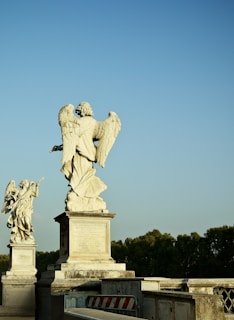 Two large stone angel statues are positioned on a bridge, with one angel holding an object while the other has its wings prominently spread. The statues are mounted on pedestals with inscriptions. The background reveals a clear blue sky and a line of trees along the horizon, contributing to a serene atmosphere.