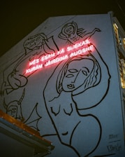 A mural on a building wall displays abstract black line drawings of human figures. Above the artwork, there is a red neon sign with text. The dark background emphasizes the stark contrast between the neon light and the murals.