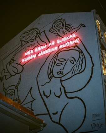 A mural on a building wall displays abstract black line drawings of human figures. Above the artwork, there is a red neon sign with text. The dark background emphasizes the stark contrast between the neon light and the murals.