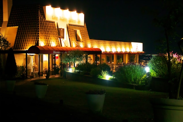A warmly lit building at night with an inviting entrance under an illuminated canopy. The structure features unique architectural elements with a tiled roof and glowing windows. Lush greenery surrounds the building, enhancing the serene nighttime ambiance.