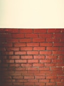 A section of a red brick wall is topped by a plain white surface. The bricks are arranged in horizontal layers with visible mortar lines, giving a textured and rustic appearance.
