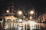 A moody cityscape at night with glowing streetlights reflecting on wet pavement.