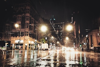 A moody nighttime cityscape with glowing lights reflecting on wet streets.