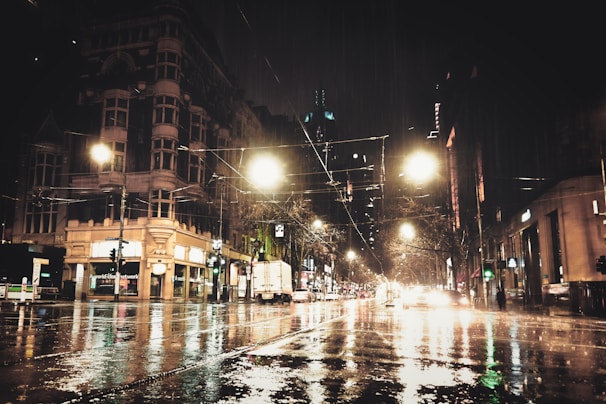 A moody nighttime cityscape with glowing lights reflecting on wet streets.