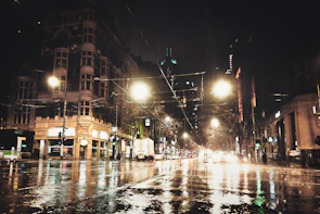 A moody cityscape at night with glowing streetlights reflecting on wet pavement.