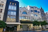 A row of stylish, multi-story buildings characterized by ornate architectural details and large windows. The prominent storefronts include Zara and David Jones/Sephora, with a modern facade. Several trees and people are seen on the bustling sidewalk in front.