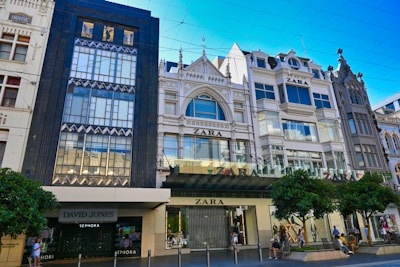 A row of stylish, multi-story buildings characterized by ornate architectural details and large windows. The prominent storefronts include Zara and David Jones/Sephora, with a modern facade. Several trees and people are seen on the bustling sidewalk in front.