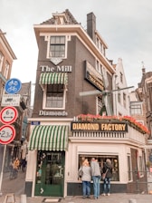 A charming, narrow building with traditional Dutch architecture houses a store named 'The Mill,' advertising diamonds. The building features dark brickwork, white accents, and a small windmill replica on its side. Green and white striped awnings cover the entrance and a side window. Above the entrance, a large sign reads 'Diamond Factory'. Bright red flowers decorate window boxes under the signs. A few people are inspecting the display window. Additionally, several road signs are visible, suggesting bicycle accessibility and height restrictions.