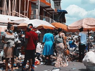 A bustling outdoor market scene with people walking and engaging with vendors. Stalls are covered with umbrellas, displaying various goods including shoes and clothing. The area is crowded and vibrant, with many individuals wearing masks.