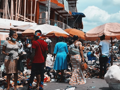 A bustling outdoor market scene with people walking and engaging with vendors. Stalls are covered with umbrellas, displaying various goods including shoes and clothing. The area is crowded and vibrant, with many individuals wearing masks.