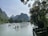 Multiple people wearing red life jackets are on bamboo rafts navigating a calm river surrounded by lush greenery and karst mountains. An umbrella on one of the rafts adds color to the scene. The sky is partly cloudy with ample sunlight illuminating the landscape. Trees overhang on the right side, framing the river view.