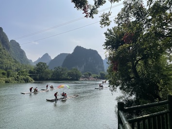 Multiple people wearing red life jackets are on bamboo rafts navigating a calm river surrounded by lush greenery and karst mountains. An umbrella on one of the rafts adds color to the scene. The sky is partly cloudy with ample sunlight illuminating the landscape. Trees overhang on the right side, framing the river view.