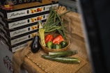 A display of fresh vegetables arranged on a rustic burlap cloth. A basket contains green onions, a red bell pepper, and two green bell peppers. Surrounding the basket are an eggplant, two cucumbers, and a bundle of asparagus. In the background, there are boxes labeled 'Australian Stone Fruit' which are stacked on top of each other.
