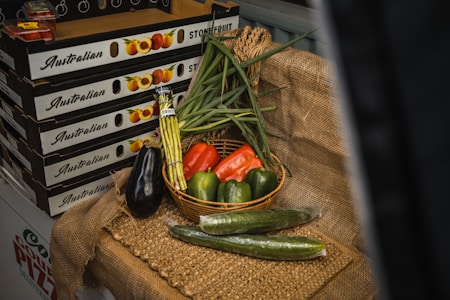 A display of fresh vegetables arranged on a rustic burlap cloth. A basket contains green onions, a red bell pepper, and two green bell peppers. Surrounding the basket are an eggplant, two cucumbers, and a bundle of asparagus. In the background, there are boxes labeled 'Australian Stone Fruit' which are stacked on top of each other.