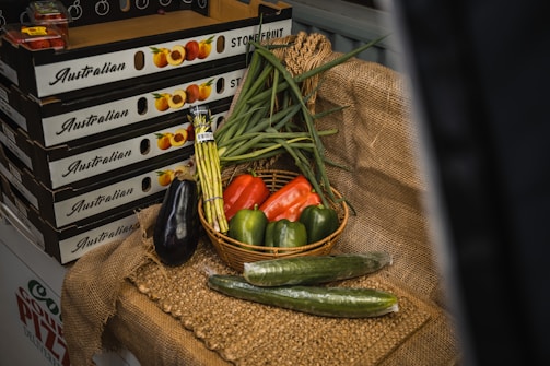 A display of fresh vegetables arranged on a rustic burlap cloth. A basket contains green onions, a red bell pepper, and two green bell peppers. Surrounding the basket are an eggplant, two cucumbers, and a bundle of asparagus. In the background, there are boxes labeled 'Australian Stone Fruit' which are stacked on top of each other.