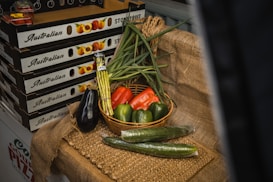 A display of fresh vegetables arranged on a rustic burlap cloth. A basket contains green onions, a red bell pepper, and two green bell peppers. Surrounding the basket are an eggplant, two cucumbers, and a bundle of asparagus. In the background, there are boxes labeled 'Australian Stone Fruit' which are stacked on top of each other.