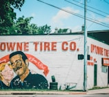 A mural is painted on the side of a building, depicting a man and a woman with a red banner in memory of two individuals. The wall includes text advertising 'Uptowne Tire Co.,' with stacked tires visible near the base.