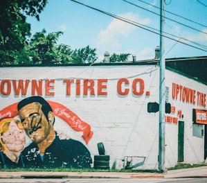 A mural is painted on the side of a building, depicting a man and a woman with a red banner in memory of two individuals. The wall includes text advertising 'Uptowne Tire Co.,' with stacked tires visible near the base.