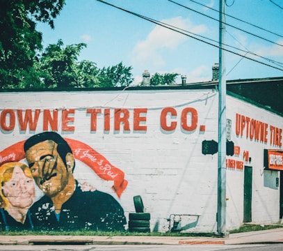 A mural is painted on the side of a building, depicting a man and a woman with a red banner in memory of two individuals. The wall includes text advertising 'Uptowne Tire Co.,' with stacked tires visible near the base.