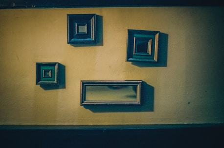 Four empty picture frames of varying sizes are mounted on a textured, yellowish wall. The frames are dark and ornate, with some having a square shape and one being rectangular. They are casting subtle shadows on the wall, adding depth to the scene.