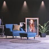 A stylish interior space with a blue upholstered armchair and matching footstool, placed on a light marble floor. A framed portrait of a woman with long red hair leans against a dark textured wall, complemented by a large green potted plant. The scene is accentuated by subtle overhead lighting and a decorative side table with a vase.