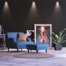 A stylish interior space with a blue upholstered armchair and matching footstool, placed on a light marble floor. A framed portrait of a woman with long red hair leans against a dark textured wall, complemented by a large green potted plant. The scene is accentuated by subtle overhead lighting and a decorative side table with a vase.