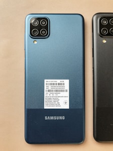 Two Samsung smartphones placed side by side on a beige surface. The phone on the left is blue and has a sticker with product details, while the one on the right is black. Both phones have a quad-camera setup in a square module at the top left corner on the back.