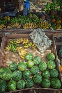 A variety of fruits including several bunches of bananas, watermelons, pineapples, and other tropical fruits are displayed on a wooden market stall. The fruits are arranged neatly with an assortment of colors and textures creating a vibrant scene. A patterned fabric bag is also visible on one side of the table.