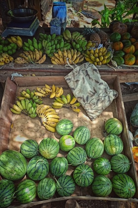 A variety of fruits including several bunches of bananas, watermelons, pineapples, and other tropical fruits are displayed on a wooden market stall. The fruits are arranged neatly with an assortment of colors and textures creating a vibrant scene. A patterned fabric bag is also visible on one side of the table.