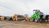 A green front loader is parked next to a stack of large cut logs on a dirt ground in an outdoor lumber yard. Wooden planks are seen neatly arranged nearby, and a small shed is in the background under a partly cloudy sky.
