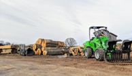 A green front loader is parked next to a stack of large cut logs on a dirt ground in an outdoor lumber yard. Wooden planks are seen neatly arranged nearby, and a small shed is in the background under a partly cloudy sky.