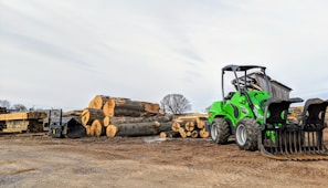 A green front loader is parked next to a stack of large cut logs on a dirt ground in an outdoor lumber yard. Wooden planks are seen neatly arranged nearby, and a small shed is in the background under a partly cloudy sky.