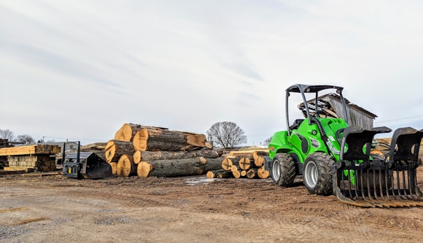 A green front loader is parked next to a stack of large cut logs on a dirt ground in an outdoor lumber yard. Wooden planks are seen neatly arranged nearby, and a small shed is in the background under a partly cloudy sky.
