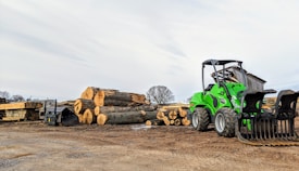 A green front loader is parked next to a stack of large cut logs on a dirt ground in an outdoor lumber yard. Wooden planks are seen neatly arranged nearby, and a small shed is in the background under a partly cloudy sky.