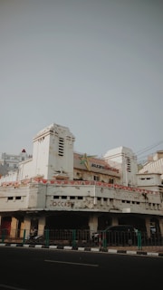 An old, weathered building with peeling paint and visible signs of age. The architecture features art deco elements, and the entrance is flanked by two cylindrical towers. Above the main entrance, a sign indicates the building might have been an insurance office. Banners with a pattern resembling national flags are draped around the facade. On the street level, a few motorbikes and cars are parked, and a green railing separates the pavement from the road.