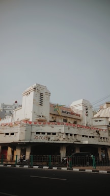 An old, weathered building with peeling paint and visible signs of age. The architecture features art deco elements, and the entrance is flanked by two cylindrical towers. Above the main entrance, a sign indicates the building might have been an insurance office. Banners with a pattern resembling national flags are draped around the facade. On the street level, a few motorbikes and cars are parked, and a green railing separates the pavement from the road.