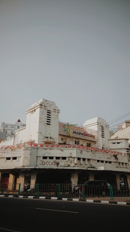 An old, weathered building with peeling paint and visible signs of age. The architecture features art deco elements, and the entrance is flanked by two cylindrical towers. Above the main entrance, a sign indicates the building might have been an insurance office. Banners with a pattern resembling national flags are draped around the facade. On the street level, a few motorbikes and cars are parked, and a green railing separates the pavement from the road.