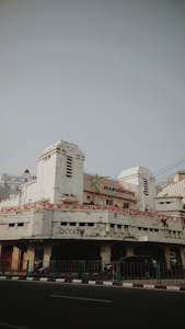 An old, weathered building with peeling paint and visible signs of age. The architecture features art deco elements, and the entrance is flanked by two cylindrical towers. Above the main entrance, a sign indicates the building might have been an insurance office. Banners with a pattern resembling national flags are draped around the facade. On the street level, a few motorbikes and cars are parked, and a green railing separates the pavement from the road.
