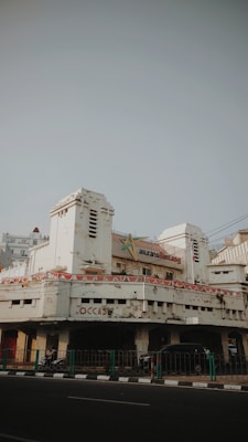 An old, weathered building with peeling paint and visible signs of age. The architecture features art deco elements, and the entrance is flanked by two cylindrical towers. Above the main entrance, a sign indicates the building might have been an insurance office. Banners with a pattern resembling national flags are draped around the facade. On the street level, a few motorbikes and cars are parked, and a green railing separates the pavement from the road.
