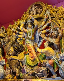 A vibrant statue depicts a multi-armed deity, adorned in ornate, colorful attire, engaged in a dynamic scene with several other figures and mythological creatures. The central figure is surrounded by intricate carvings and details, with expressions of strength and leadership.