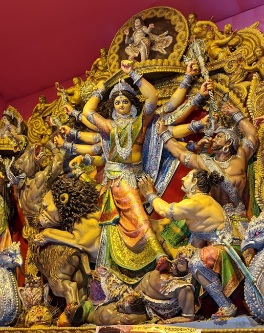 A vibrant statue depicts a multi-armed deity, adorned in ornate, colorful attire, engaged in a dynamic scene with several other figures and mythological creatures. The central figure is surrounded by intricate carvings and details, with expressions of strength and leadership.