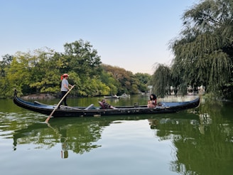 A gondola is floating on a calm, green-tinted body of water surrounded by lush greenery. A person dressed in a striped shirt is standing and rowing the gondola, while another person is seated inside, enjoying the ride. Trees are reflected in the water, adding to the tranquil ambiance.