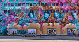 A large and colorful street mural covers a building's facade featuring portraits of historical figures intertwined with vibrant abstract designs and architectural elements. The mural uses a palette of pink, teal, and gold, showcasing a mix of surreal and cubist styles with intricate details. Below the mural, there are pedestrians and a parked car.