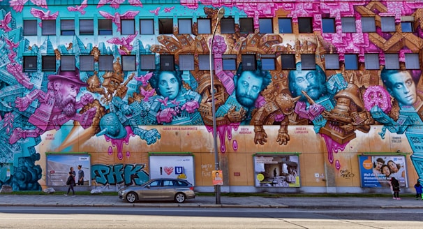 A large and colorful street mural covers a building's facade featuring portraits of historical figures intertwined with vibrant abstract designs and architectural elements. The mural uses a palette of pink, teal, and gold, showcasing a mix of surreal and cubist styles with intricate details. Below the mural, there are pedestrians and a parked car.