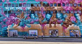 A large and colorful street mural covers a building's facade featuring portraits of historical figures intertwined with vibrant abstract designs and architectural elements. The mural uses a palette of pink, teal, and gold, showcasing a mix of surreal and cubist styles with intricate details. Below the mural, there are pedestrians and a parked car.